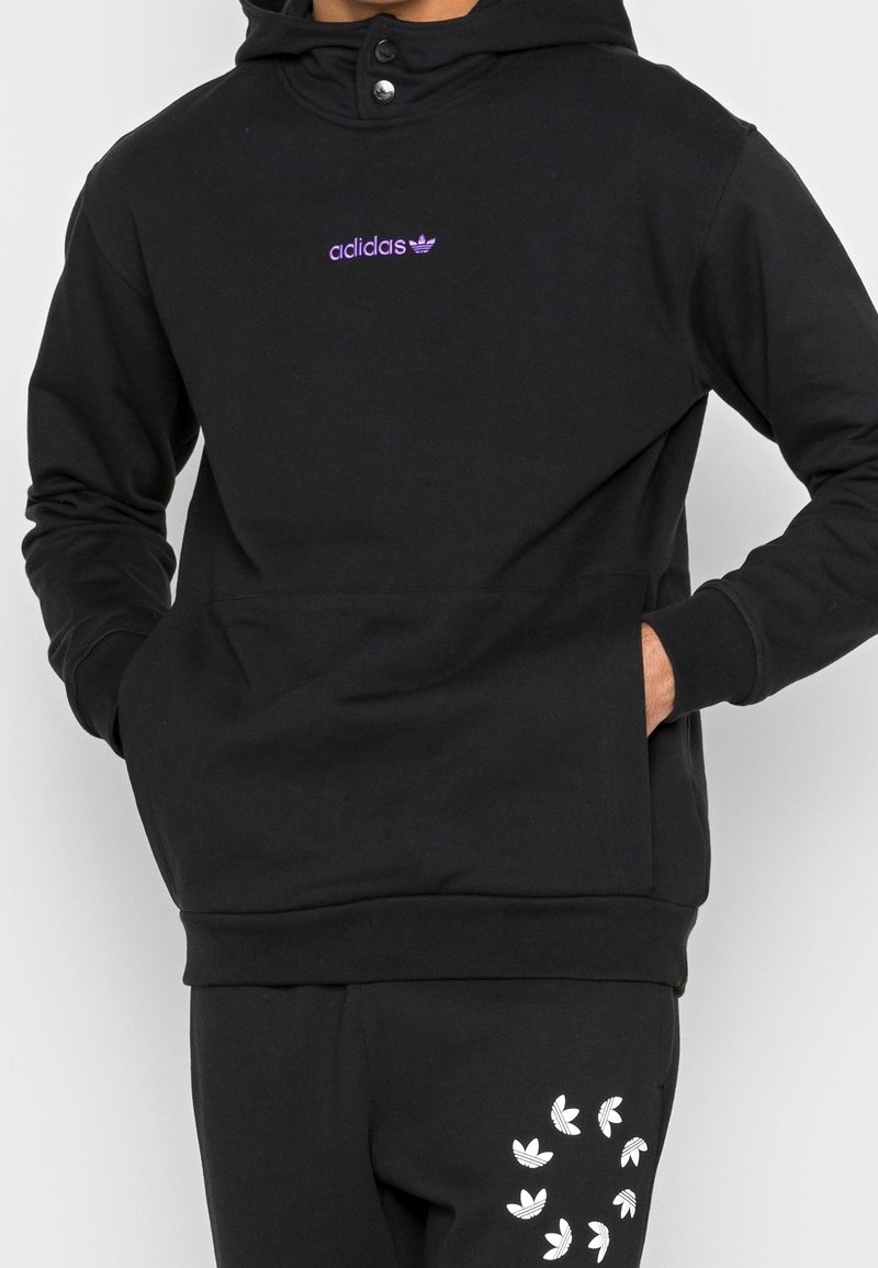 Svart huvtröja med känguruficka, prydd med en lila broderad logotyp. Slät textur, justerbar dragsko och tryckknappsstängning.