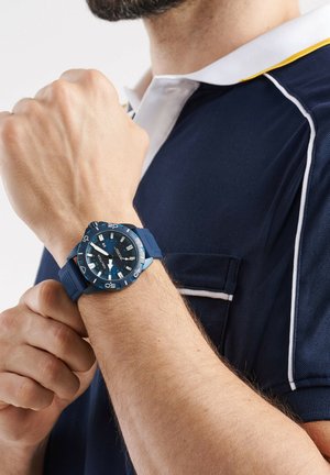 Montre analogique bleue avec un cadran rond, un bracelet en caoutchouc et des aiguilles lumineuses. Portée sur un poignet avec une texture de peau visible, contre une chemise col marin.