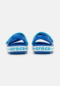 Crocs albastre cu o suprafață texturată, curea la călcâi și design deschis la spate. Prezintă logo-ul "crocs" în albastru deschis pe o talpă albă.