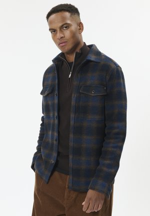 Kostkovaná modro-hnědá košile s bundovými vlastnostmi z měkké látky, která disponuje dvěma náprsními kapsami, zapínáním na knoflíky a límečkem, nošená přes tmavý svetr.