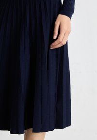 Navy pleated skirt made of textured fabric, featuring vertical pleats and a ribbed waistband, shown with a hand placed on the fabric.