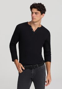 Camicia henley nera a maniche lunghe con un design testurizzato, dotata di una morbidissima abbottonatura e vestibilità rilassata, abbinata a jeans in denim grigi e una cintura nera.