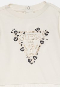 Kreemjas puuvillane kleit, millel on sädeleva kolmnurkse logo tekstiga "GUESS?" kuldse ja musta värviga, kaunistatud südame ja leopardimustriga aksendidega.