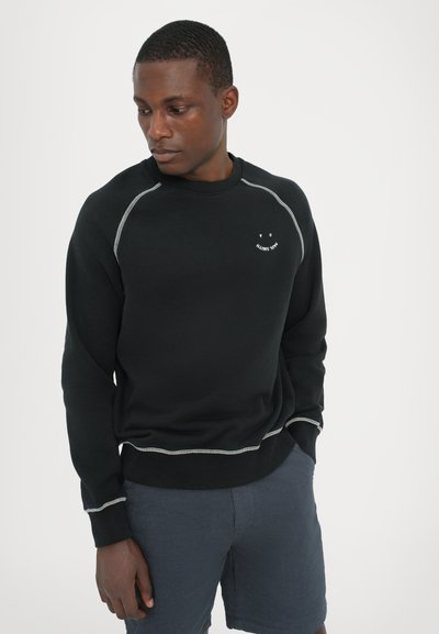 Svart sweatshirt med kontrasterande sömmar, rund halsringning och leende-logotyp på bröstet. Mjuk tyg; avslappnad passform. Matchas med blå shorts.