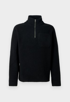Mustan fleece-neuleen, jossa on korkea kaulus ja puolipitkä vetoketju. Vasemmassa rinnassa on läpällinen tasku ja pintarakenteessa on tekstuuri.