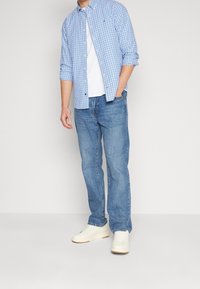 Blau-weiße karierte Button-Down-Hemd über einem weißen T-Shirt, kombiniert mit blauen Jeans und weißen Sneakers. Lässiger Look mit hochgekrempelten Ärmeln.