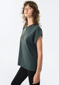 Zielona koszula z krótkim rękawem i dekoltem w kształcie litery V, o luźnym kroju, z fakturowanym napisem z przodu. Noszona z czarnymi legginsami.