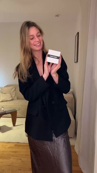 Blazer noir surdimensionné, jupe en satin argenté, tenant une petite boîte blanche avec une étiquette noire portant l'inscription "Calvin Klein". Cadre intérieur avec canapé en arrière-plan.