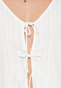 Λευκή μπλούζα με βαθύ V ντεκολτέ και κάθετη ριγέ υφή, στερεωμένη με δύο λευκούς δεσμούς στο λαιμό. Μαλακό ύφασμα με ελαφριά εμφάνιση.