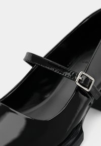Chaussure Mary Jane en cuir verni noir avec une bride à boucle argentée et un talon bas sur fond blanc.