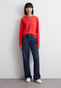 Rød langærmet skjorte parret med højtaljede, løstsiddende mørkeblå jeans og hvide sneakers, der skaber et afslappet outfit. Enkel sølvhalskæde som pynt.