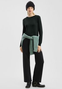 Zwarte langarmshirt, zwarte wijde broek en een lichtgroene gebreide omslag om de taille. Zwarte beanie en enkellaarsjes met hardware-details.