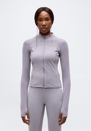 Frau trägt eine eng anliegende hellgraue Sportjacke mit Reißverschluss und passende Leggings, steht vor einem schlichten weißen Hintergrund.