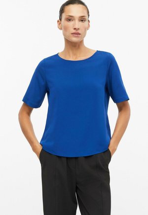 Frau mit dunklem zurückgebundenem Haar, die eine schlichte, leuchtend blaue, kurzärmelige Bluse und schwarze Hosen trägt, mit Händen in den Taschen, vor weißem Hintergrund.