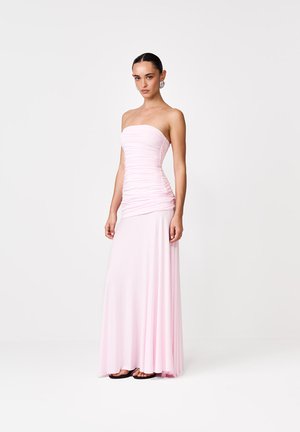 Naine, kes kannab rihmadeta, voltidega heleroosat põrandapikkust kleiti ja rõngakujulisi kõrvarõngaid, seisab tavalise valge tausta ees.