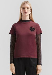 Burgunderrotes T-Shirt mit kurzen Ärmeln und rundem Halsausschnitt, das ein bedrucktes Herzdesign auf der Brust aufweist, darüber geschichtet mit einem schwarzen, Netz-Löwenmuster Langarmshirt.