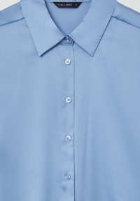 Camicia azzurro chiaro a maniche corte realizzata in tessuto morbido, con colletto a punta e sei bottoni lungo la parte anteriore.