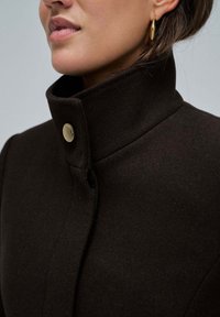 Abrigo de lana marrón con un collar alto, que presenta un detalle de botón dorado cerca del escote. Textura suave y diseño minimalista.