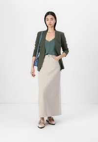 Olijfgroen blazer over een teal top, gecombineerd met een crèmekleurige maxirok met verticale strepen en beige instappers met decoratieve hardware.