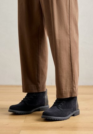 Botas de ante negras hasta el tobillo con un acabado texturizado, con cordones y una suela robusta, combinadas con pantalones marrones de pierna ancha, sobre un suelo de madera.