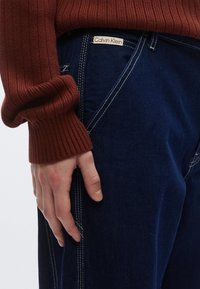 Vaqueros de mezclilla azul con costuras blancas y una etiqueta de Calvin Klein. Un suéter de canalé color burdeos con los puños enrollados es visible, cubriendo parte de una mano.