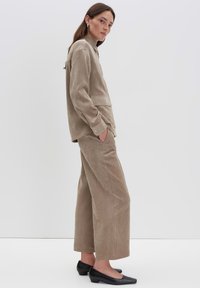 Completo in cotone beige con camicia colletto e pantaloni a gamba larga; tessuto strutturato con tasche laterali e scarpe nere piatte.
