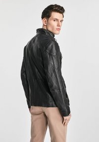 Czarna skórzana kurtka z wysokim kołnierzem, dopasowanym krojem i subtelnymi detalami przeszycia, noszona na beżowych spodniach. Gładka faktura.