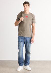 Tricou din bumbac verde măsliniu cu un patch logo roșu, purtat cu blugi deschiși la culoare și adidași albi, stând pe un podea neutră.