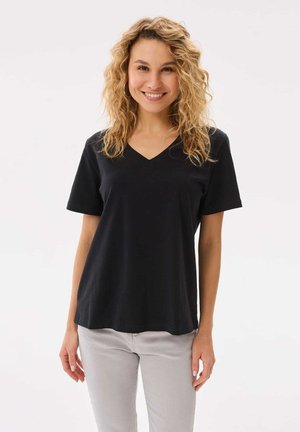 Uśmiechnięta kobieta o kręconych blond włosach, ubrana w czarną koszulkę z dekoltem w serek i jasnoszare spodnie, stojąca na białym tle.