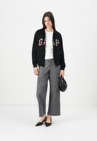 Black zip-up hoodie with floral "GAP" lettering, white fitted t-shirt, grey wide-leg cargo pants, and black pointed-toe heels. Small black bag.