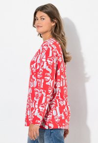 Rotes Langarm-T-Shirt mit weißem Text und Gesichtern in einem wiederkehrenden Muster. Rundhalsausschnitt und lockere Passform. Weicher Stoff.