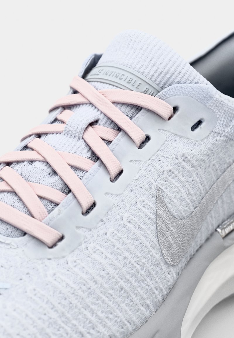 Scarpa da corsa grigia chiara con tomaia in maglia texture, lacci rosa e logo Nike riflettente argentato sul lato.