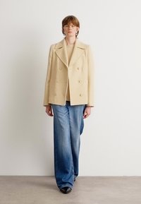 Béžový dvojradový kabát s zaoblenými klopami, v kombinácii so svetlomodrými džínmi s širokými nohavicami. Hladká textúra látky a veľké gombíky.