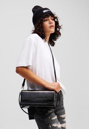 Borsa crossbody in finta pelle nera lucida con logo embossato, forma rettangolare, tracolla staccabile, abbinata a una camicia bianca e jeans strappati.