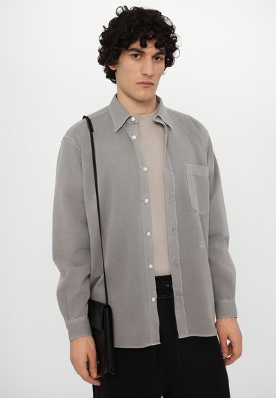 Chemise boutonnée gris clair à manches longues et avec une poche poitrine. Associée à une chemise beige en dessous et portée avec un sac bandoulière noir.