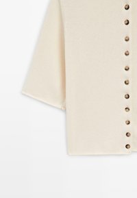 Krémově zbarvený pletený kardigan s krátkými rukávy a svislým řaděním malých hnědých knoflíků vpředu.