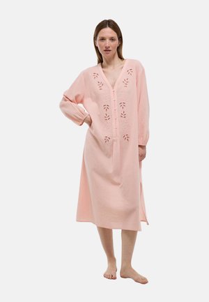 Femme vêtue d'une longue robe rose ample avec un devant boutonné et des motifs floraux découpés, debout pieds nus sur un fond blanc uni.