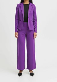 Kvinna klädd i en lila kavaj och vida byxor, matchad med en svart topp och svarta spetsskor mot en vit bakgrund.