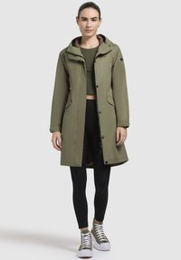 Olivgrön vadderad parkas med huva, framknapp och dragkedjestängning, samt foder i leopardmönster. Bärs över svarta leggings och gröna sneakers.