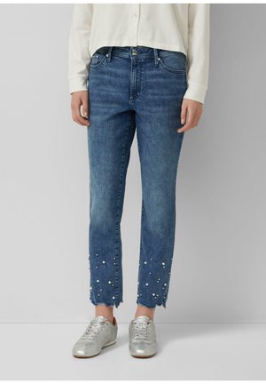 Niebieskie dżinsy z denimem z postrzępionymi krawędziami i rozsypanymi białymi perłami w okolicy kostek, noszone z srebrnymi trampkami i białą bluzką z długim rękawem.