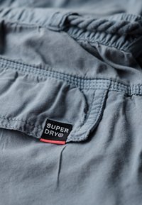 Szara tkanina o miękkiej strukturze, z elastycznym pasem i widoczną czarną oraz czerwoną metką "SUPER DRY". Obecne są również szwy dekoracyjne.