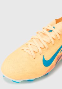 Jasnopomarańczowe korki piłkarskie Nike z niebieskim logo swoosh, sznurowadłami i niebieskimi wkrętami na białym tle.