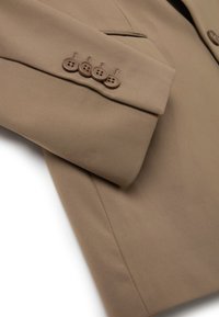Manica di blazer beige con quattro bottoni marroni. Tessuto morbido con texture liscia e design sartoriale. Cuciture dettagliate visibili vicino ai bottoni.