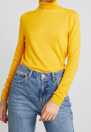Personne portant un pull jaune à col roulé et manches longues, rentré dans un jean bleu taille haute, sur un fond uni.