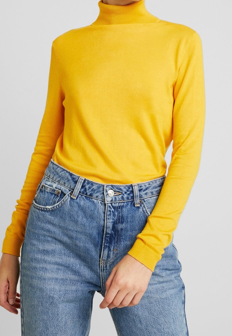 Personne portant un pull jaune à col roulé et manches longues, rentré dans un jean bleu taille haute, sur un fond uni.