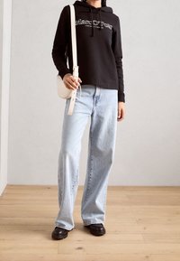 Sudadera negra con capucha y logo blanco, jeans de pierna ancha azul claro, bolso cruzado beige y zapatos gruesos marrón oscuro sobre un suelo de madera.