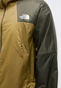 Dvojfarebná bunda v olivovo zelenej a tmavo zelenej farbe s vreckom na zips a bielym logom The North Face na hrudi.