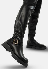 Botas altas de cuero negro con suelas gruesas, correa de hebilla dorada cerca del tobillo y cremallera lateral, mostradas en posición de pie sobre un fondo blanco.