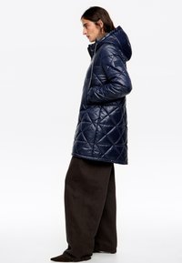 Námornícko modrá prešívaná páperová bunda s kapucňou, so zipsom, textúrovanou látkou a bočnými vreckami. V kombinácii so širokými hnedými nohavicami.