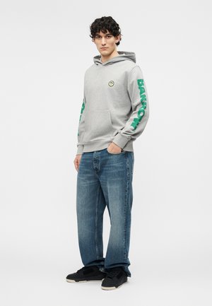 Jonge man die een grijze hoodie draagt met groene tekst op de mouwen en een blauwe spijkerbroek, staand met één hand in zijn zak en zwarte sneakers.